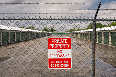 No trespassing sign on a barbed wire fence with secure storage units in the background.
